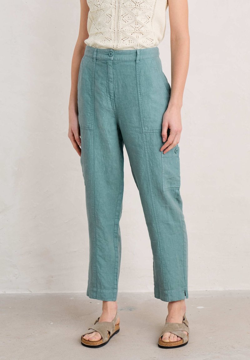 Femme portant un pantalon en lin taille haute couleur sarcelle avec poches avant et latérales et des sandales beiges à bout ouvert, debout contre un mur uni.