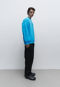 Blauwe oversized sweatshirt, zwarte broek en zwarte sportschoenen. Gladde stof, relaxte pasvorm en opvallende manchetten. Krullend haar in een natuurlijke stijl.