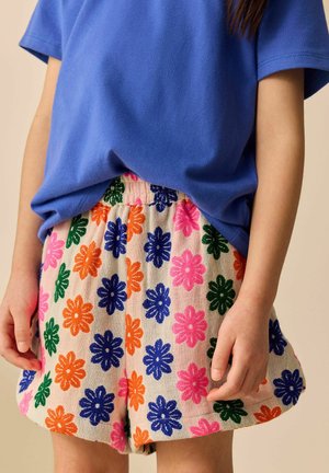 Kind draagt een blauw shirt met korte mouwen en beige korte broek met kleurrijke roze, oranje, blauwe en groene bloemenpatronen.