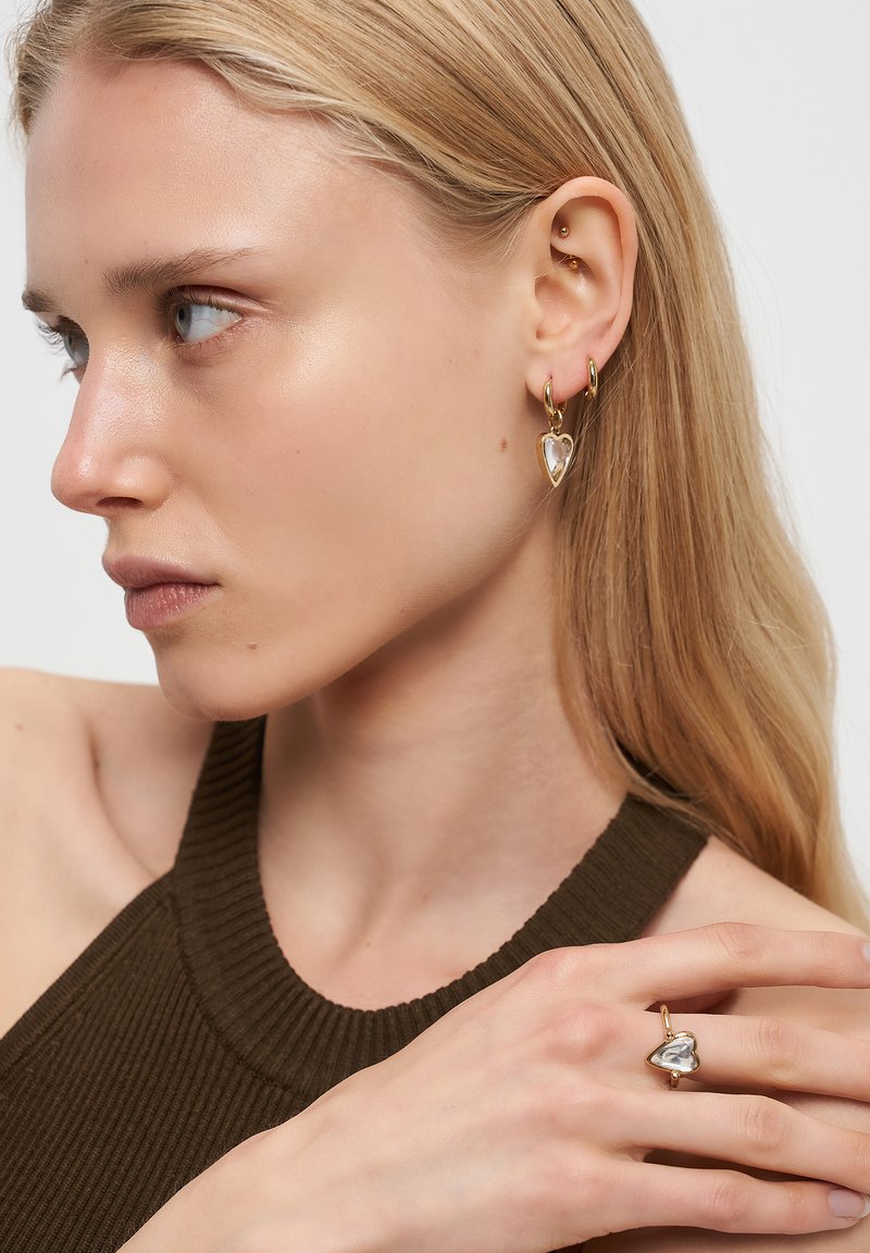 Blond kvinna i en mörk ärmlös top, som visar upp guldfärgade hjärtformade örhängen och en matchande hjärtformad ring.