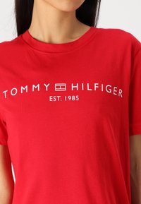 Red cotton t-shirt featuring white text "TOMMY HILFIGER EST. 1985" with a logo design. Short sleeves and crew neck style.