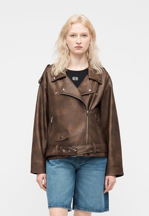 Blonde Person trägt eine braune Lederjacke mit einem Taillengürtel und blaue Jeansshorts und steht vor einem schlichten Hintergrund.