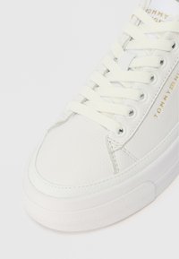 Witte leren sneaker met witte veters, gestructureerde zool en gouden "Tommy Hilfiger" branding aan de zijkant en tong.