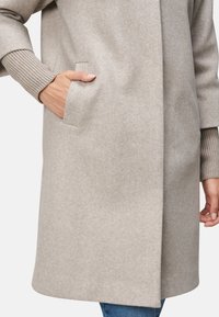 Manteau en mélange de laine gris clair avec des poignets en tricot côtelé et des poches latérales. Coupe droite, design mi-long, texture lisse, détails minimalistes.