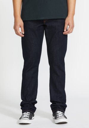 Personne portant un jean bleu foncé et des baskets basses noires avec des lacets blancs, debout devant un fond uni.