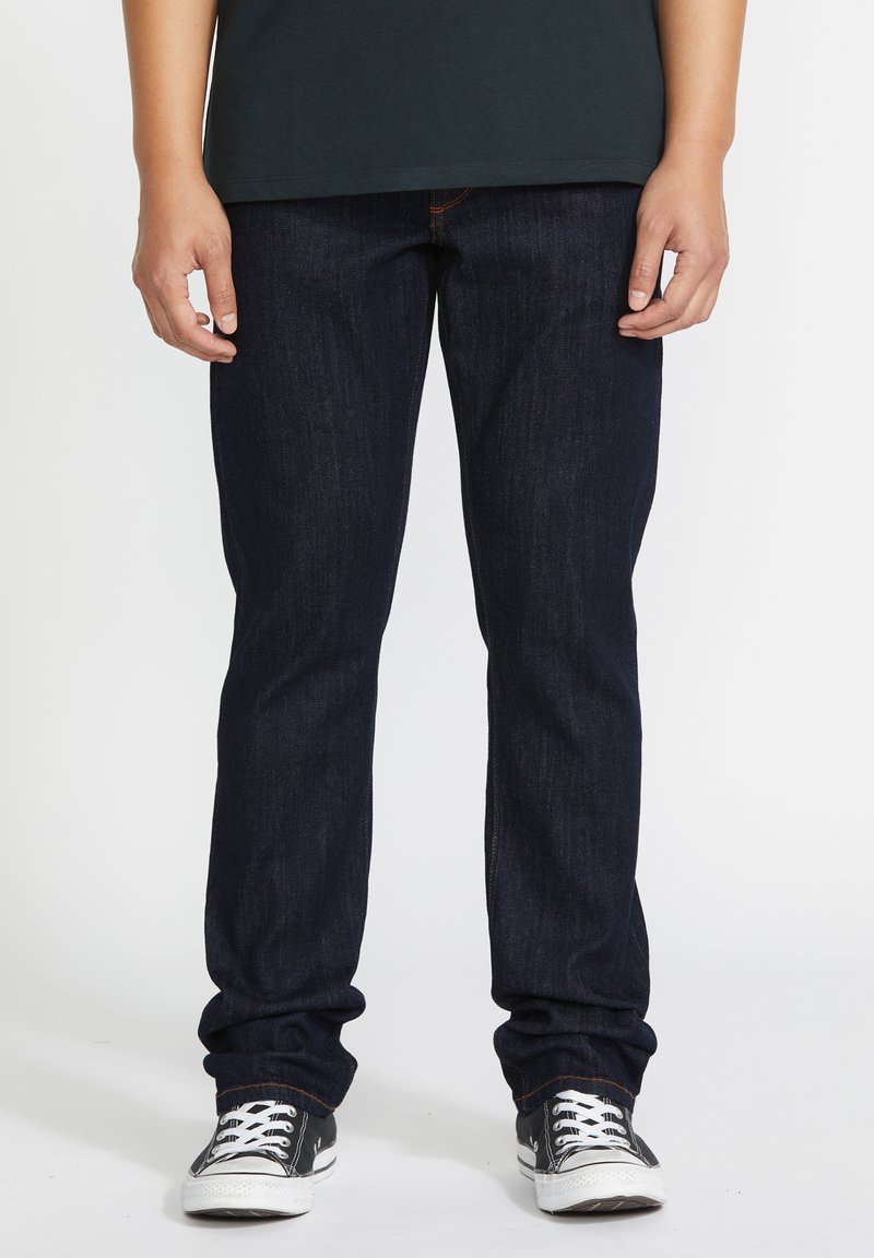 Personne portant un jean bleu foncé et des baskets basses noires avec des lacets blancs, debout devant un fond uni.