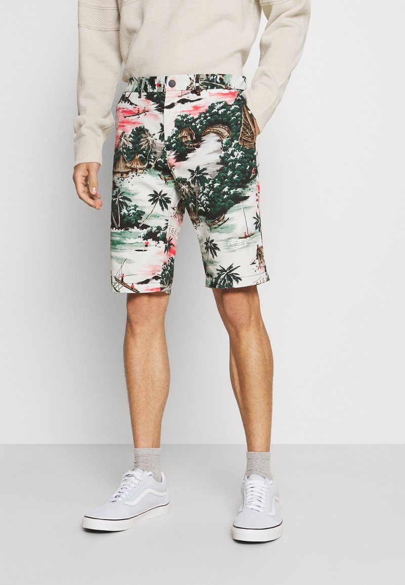 Homme portant des baskets blanches, des chaussettes gris clair, un short imprimé à thème tropical, et un pull beige à manches longues avec une main dans la poche.