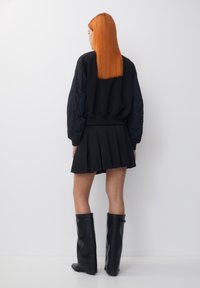 Blouson aviateur noir avec manches rembourrées, associé à une jupe noire plissée et des bottes en cuir noires montantes jusqu'aux genoux. Cheveux longs et raides de couleur orange visibles.