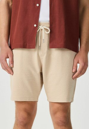 Beige strukturierte Kordelzug-Shorts, getragen mit einem weißen gerippten Tanktop und einem ungebundenen, rostfarbenen Hemd, Hände entspannt an den Seiten.