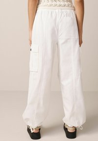 Pantalons cargo blancs avec une taille élastique, des poches latérales et des poignets à cordon de serrage. Fabriqués en tissu léger avec une texture lisse.