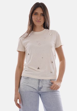 Donna che indossa una T-shirt beige a maniche corte con applicazioni argentate sparse e jeans azzurri con dettagli sottili di strass.