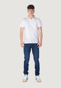 Camisa polo blanca con cuello beis, jeans ajustados azules y zapatillas blancas con detalles verdes, de pie contra un fondo neutro.