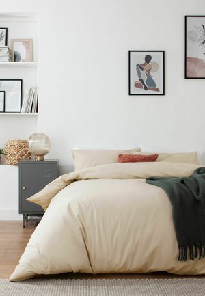 Beiges Bett mit Kissen und dunkelgrüner Decke neben einem grauen Nachttisch mit Lampe und Korb, mit abstrakter Kunst und Regalen an weißen Wänden.