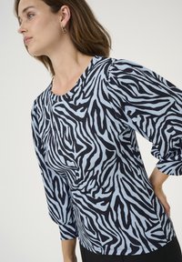 Sieviete valkā zilas un melnas zebras svītras ar pūkainām garām piedurknēm, stāvot ar roku uz gurniem pret vienkāršu fonu.