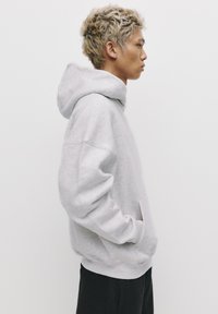 Szary polarowy hoodie z kieszenią kangurzą, w oversize'owym fasonie, z kapturem ze sznurkiem. Posiada ściągacze przy mankietach i dolnej krawędzi. Noszony z czarnymi spodniami.