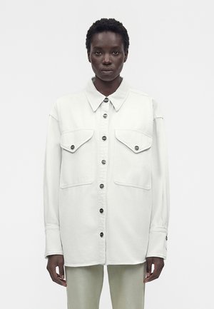 Personne à la peau foncée portant une chemise blanche surdimensionnée à boutons avec deux grandes poches poitrine, debout devant un fond clair uni.