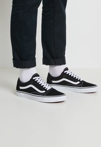 Baskets en daim noir avec des accents en cuir blanc, présentant un design bas classique, des lacets blancs et des semelles en caoutchouc, portées avec un pantalon noir retroussé.