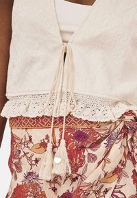 Top tejido crema con textura floral, un borde de encaje con forma de escallón y un cierre con cordón y borlas. Combinado con una falda de patrón floral en tonos cálidos.
