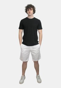 T-shirt nera a maniche corte abbinata a pantaloni bianchi. Modello che indossa scarpe da ginnastica bianche, in piedi diritto con le mani in tasca, sfondo neutro.
