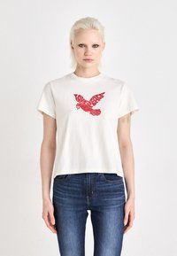 Blondína v bielom tričku s červenou grafikou vtáka a textom "LOVE PEACE", kombinovaná s modrými džínami, stojí proti bielemu pozadiu.