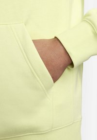 Felpa gialla chiaro con tasca a marsupio e polsini a coste. Il tessuto appare morbido e dalla texture particolare. Una mano è poggiata nella tasca.