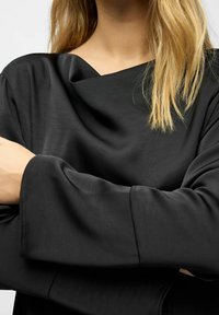 Schwarzes, in glattem Stoff drapiertes Oberteil mit weitem Ausschnitt und langen Ärmeln, in lässiger Passform und minimaler Detailgestaltung.