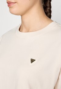 Lichtbeige t-shirt met korte mouwen met een driehoekig logo patch met een gouden accent aan de linkerkant van de borst. Zachte stoftextuur.