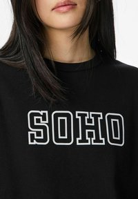 Moteris su tiesiais tamsiais plaukais, vilkinti juodą sportinį megztinį su baltais kontūrais raidžių "SOHO" ant krūtinės.