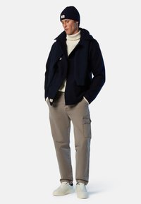 Cappotto navy a doppio petto sopra un dolcevita crema, con pantaloni cargo kaki e sneakers bianche. Il look è completato da un berretto di lana navy.