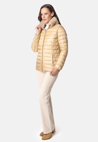 Chaqueta de abrigo dorada con un cuello forrado de piel, textura suave, cierre con cremallera en la parte frontal, diseño acolchado, combinada con pantalones de campana de color crema y botas beige.