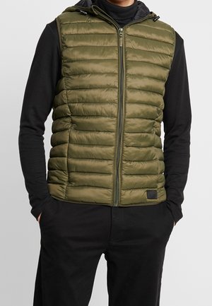 Homme portant un gilet matelassé sans manches de couleur vert olive par-dessus une chemise noire à manches longues et un pantalon noir, les mains dans les poches.