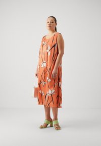 Oranje bloemenjurk met witte bloemen en zwarte lijnpatronen; gecombineerd met groene blokhak sandalen en een witte handtas.