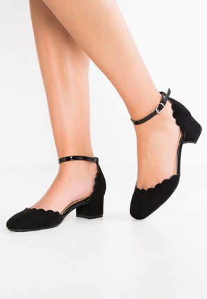 Zwarte suède lage hakken schoenen met een scalloped randontwerp en enkelriem. Rond neusvorm en textuurafwerking.