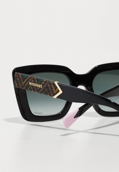 Černé oversized sluneční brýle s gradientovými čočkami. Mají vzorovanou nohu se zlatými akcenty a růžovým zakončením. Designové logo je viditelné na noze.