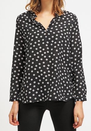 Blouse noire à manches longues avec motif de points circulaires blancs, portée non rentrée sur un pantalon noir par une personne aux cheveux mi-longs.
