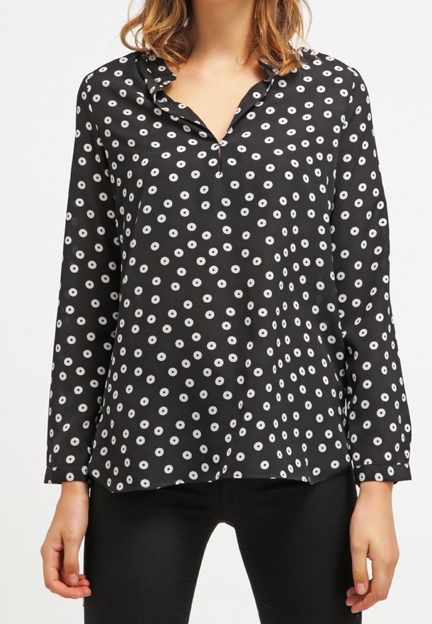 Blouse noire à manches longues avec motif de points circulaires blancs, portée non rentrée sur un pantalon noir par une personne aux cheveux mi-longs.