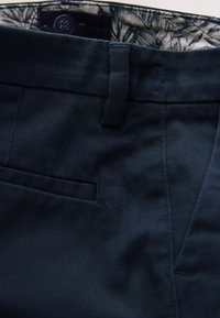 Gros plan d'un pantalon bleu foncé montrant un passant de ceinture, une poche latérale, un bouton et une ceinture intérieure à motifs.