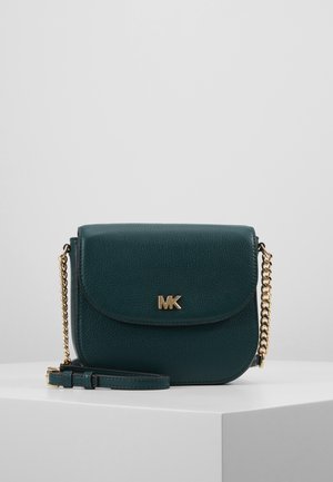 Sac bandoulière en cuir vert foncé avec une chaîne dorée et le logo "MK" sur le devant, présenté sur un socle blanc sur fond gris.
