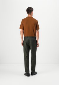 Smeđa kratka polo majica sa kratkim rukavima u paru s tamnozelenim hlačama. Outfit se ističe teksturiranom tkaninom i uskim krojem. Crne cipele upotpunjuju izgled.
