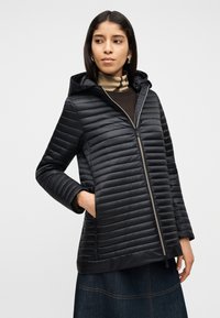 Γυναίκα που φοράει μαύρο πλεκτό μπουφάν με κουκούλα και φερμουάρ, τα χέρια στις τσέπες, κοιτάζοντας ελαφρώς προς τα πλάγια μπροστά σε απλό φόντο.