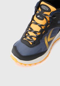 Buty sportowe z oddychającą siateczką w granatowym i czarnym kolorze, z żółtymi akcentami i szarymi sznurówkami. Posiadają wytrzymałą gumową podeszwę z pomarańczowym bieżnikiem.