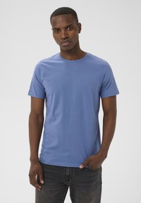 Homme aux cheveux courts portant un t-shirt bleu uni à manches courtes et col rond, ainsi qu'un jean foncé, debout avec une main dans la poche.