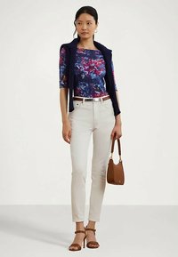 Naine seisab, seljas lillemuster pluus, valged püksid, tumesinine kampsun õlgadel, pruunid kontsad ja hoiab pruunist käekotti.