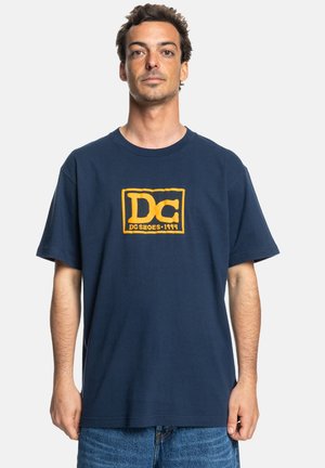 Navyblauwe katoenen t-shirt met een gele "DC"-logo binnen een rechthoekige omtrek, korte mouwen en een relaxte pasvorm.