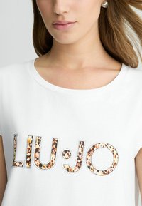 Balta trumpa rankovėmis marškinėliai, kurių dizainas pasižymi „LIU·JO“ logotipu leopardų raštu su sidabriniais akcentais. Minkšta medžiaga ir apvali iškirptė.