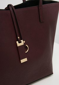 Borsa a spalla in faux leather color marrone scuro con una texture liscia, dotata di un accento in metallo dorato e un'etichetta con tracolla removibile.