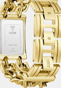 Guld stainless steel ur med en bred kædearmbånd, firkantet urskive og indgraveret brandlogo. Har polerede og børstede metalelementer.