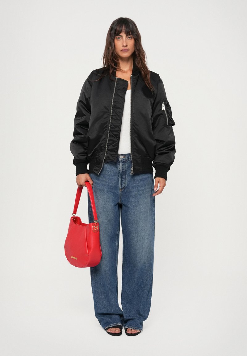 Casaco bomber preto com fecho à frente, punhos canelados e um bolso lateral, combinado com jeans de perna larga e uma bolsa vermelha com um detalhe em ouro.