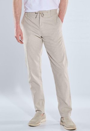 Mann trägt hellbeige Hose mit Kordelzug und passende Slipper, steht mit einer Hand in der Tasche vor einfarbigem Hintergrund.