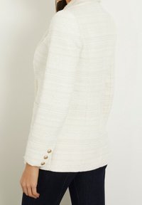 Biała marynarka z tweedu o fakturowanym wzorze, z kołnierzem i złotymi guzikami na rękawach. Dopasowany krój, o długości do połowy uda.